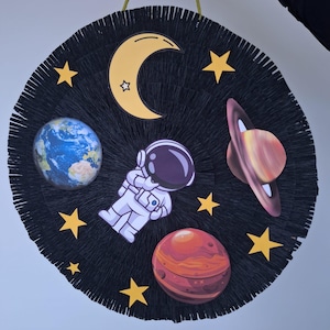 May include: A circular, black, fringed decoration with a space theme. It features a crescent moon, stars, planets, and an astronaut. The planets include Earth, Mars, and Saturn. It is suspended by a gold cord.
