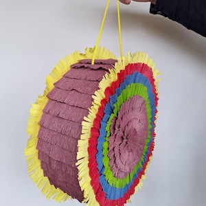 May include: A colourful, round piñata with a yellow hanging string. The piñata features layers of purple, red, blue, green, and yellow fringed paper. The piñata is held by a person.