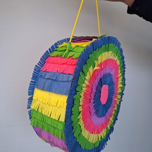 Könnte beinhalten: Eine bunte, runde Piñata mit Regenbogenmuster. Die Piñata besteht aus geschichtetem Krepppapier in Blau-, Grün-, Gelb-, Rosa- und Lilatönen. Sie hat eine gelbe Schnur zum Aufhängen.