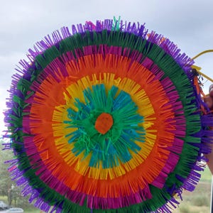 May include: A circular piñata featuring concentric rings of fringed paper in vibrant colours including purple, green, orange, yellow, and blue. The design radiates from a central orange circle, creating a layered, textured appearance with thin paper strips.