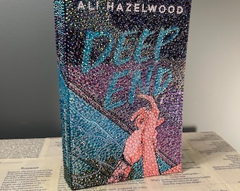 May include: A book cover with a glittery, iridescent design. The title "Deep End" is written in white letters. The author's name, "Ali Hazelwood", is written in white letters above the title. The book cover features a hand reaching out from the water.
