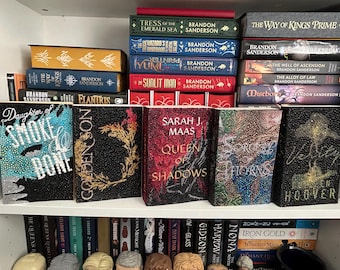 May include: A bookshelf with a variety of fantasy novels, including "Daughter of Smoke & Bone", "Elantris", "Queen of Shadows", "Sorcery of Thorns", and "Vow of Thieves".