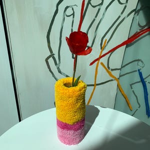Pode incluir: Uma tulipa vermelha num vaso cilíndrico. O vaso é coberto por um tecido texturizado e colorido, com secções amarelas, rosa e rosa claro. O vaso está sobre uma superfície branca, com uma pintura no fundo.