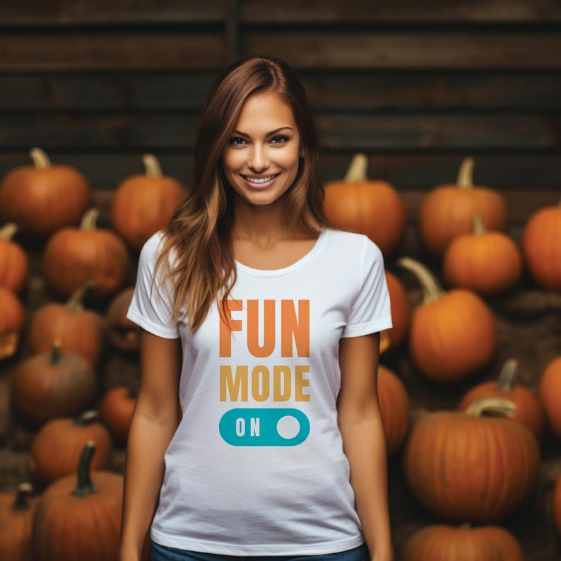 May include: A white t-shirt with the words "FUN MODE ON" in orange, yellow, and teal. The shirt is being worn in front of a backdrop of pumpkins. The shirt is a crew neck style.