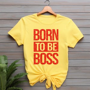May include: A yellow t-shirt with the text "BORN TO BE BOSS" in red block letters. The shirt is knotted at the waist and hangs on a wooden hanger. The background is a wooden wall with green plants.