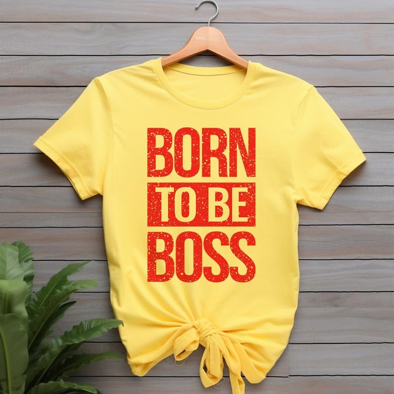 May include: A yellow t-shirt with the text "BORN TO BE BOSS" in red block letters. The shirt is knotted at the waist and hangs on a wooden hanger. The background is a wooden wall with green plants.