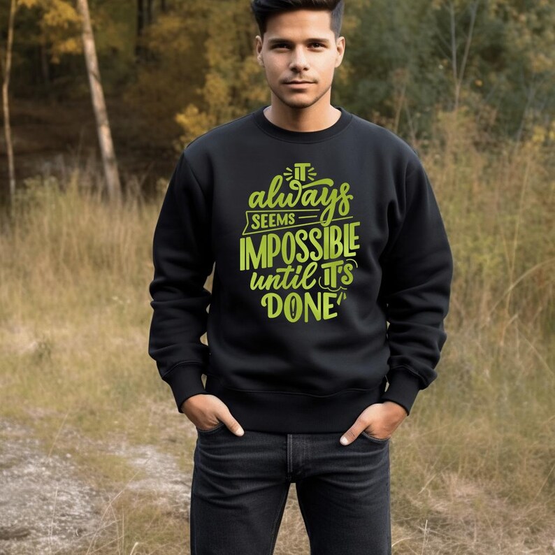 May include: Black sweatshirt with the text "It always seems impossible until it's done" in bright green. The sweatshirt is a crew neck style and is being worn by a person in a natural outdoor setting.