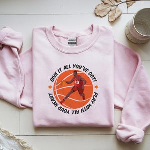 May include: A light pink sweatshirt featuring a basketball graphic with a player in red and the text "GIVE IT ALL YOU'VE GOT! PLAY WITH ALL YOUR HEART". The sweatshirt has long sleeves and a crew neck.
