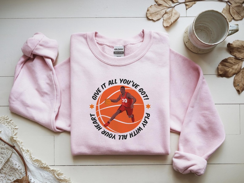 May include: A light pink sweatshirt featuring a basketball graphic with a player in red and the text "GIVE IT ALL YOU'VE GOT! PLAY WITH ALL YOUR HEART". The sweatshirt has long sleeves and a crew neck.