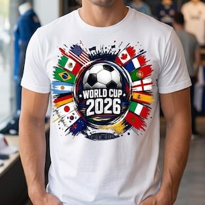 May include: A white short-sleeve t-shirt featuring a central graphic of a soccer ball surrounded by various national flags. The design includes the text "WORLD CUP 2026" in bold white lettering set against a dark circular background with colorful paint-like splashes.