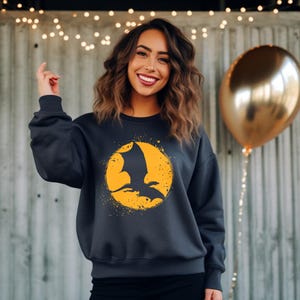 May include: Dark gray sweatshirt featuring a yellow circle with a black bat silhouette. The bat is in flight against a textured yellow background. The sweatshirt has a crew neck and long sleeves.