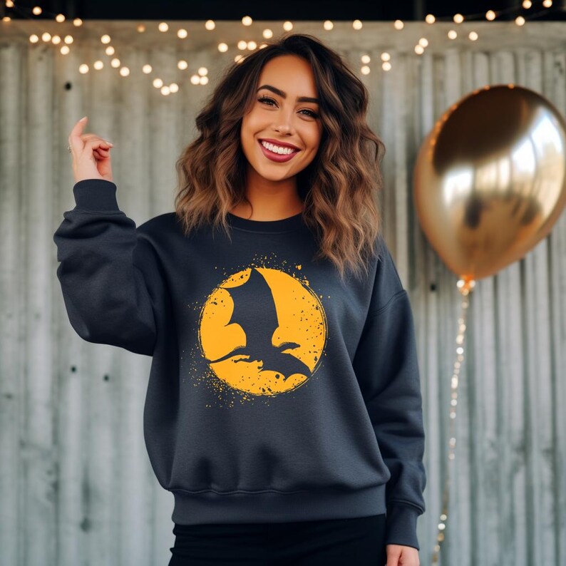May include: Dark gray sweatshirt featuring a yellow circle with a black bat silhouette. The bat is in flight against a textured yellow background. The sweatshirt has a crew neck and long sleeves.