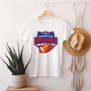 May include: White t-shirt featuring a graphic of an American football inside a blue and red shield with the words "AMERICAN FOOTBALL". The football is orange and white. The shirt is hanging on a wooden hanger.