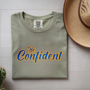 May include: A sage green t-shirt with the words "Be Confident" in blue and yellow script. The shirt is folded neatly on a light wood surface, with a straw hat and a cactus nearby.