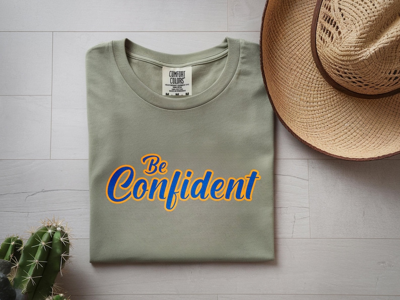 May include: A sage green t-shirt with the words "Be Confident" in blue and yellow script. The shirt is folded neatly on a light wood surface, with a straw hat and a cactus nearby.
