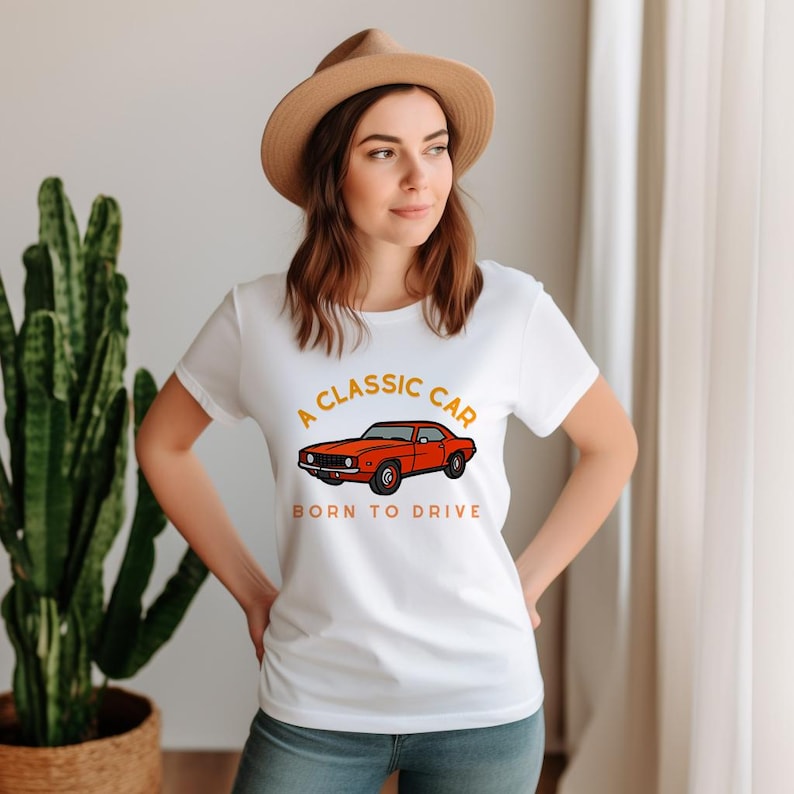May include: White t-shirt with a classic car graphic in orange and black. The text on the shirt reads "A CLASSIC CAR" above the car image and "BORN TO DRIVE" below it. The shirt is worn by a person wearing a brown hat.