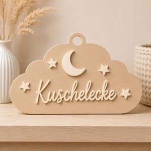Könnte beinhalten: Ein helles Holzschild in Wolkenform mit einem runden Loch an der Oberseite zum Aufhängen. Das Schild ist mit einem erhabenen Halbmond, vier kleinen Sternen und dem Schriftzug "Kuschelecke" in Schreibschrift auf der Vorderseite versehen.