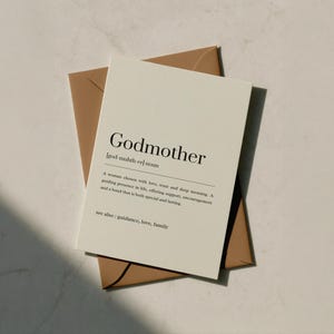 Peut inclure: Une carte de vœux crème avec le mot "Godmother" imprimé en noir, ainsi qu'une définition. La carte est posée sur une enveloppe marron. Le texte "see also: guidance, love, family" est en bas.