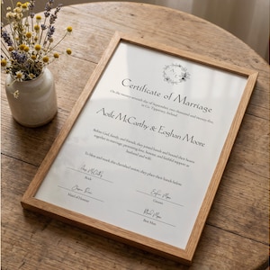 Op de afbeelding: Een ingelijste huwelijksakte met een licht houten frame. De akte heeft zwarte tekst op een witte achtergrond, met de namen Aoife McCarthy & Eoghan Moore. Een kleine vaas met gedroogde bloemen staat links van het frame.