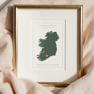Könnte beinhalten: Gerahmter Druck mit einer dunkelgrünen Karte von Irland und einem roten Herz, das einen Ort markiert. Der Druck enthält Text oben und unten, alles in einem goldfarbenen Rahmen. Der Hintergrund ist ein hellbeiger Stoff.