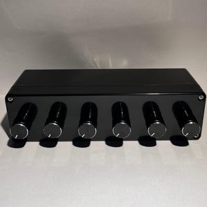 Puede incluir: Dispositivo electrónico negro con seis perillas plateadas. El dispositivo rectangular lleva la inscripción "Idle Works" en la esquina superior izquierda. Las perillas están espaciadas uniformemente en la parte delantera.