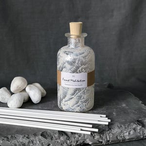 May include: A glass diffuser bottle with a wooden stopper, displaying a silver and white textured pattern. The label reads "Forest Meditation." White diffuser sticks and smooth white stones are arranged nearby. A decorative item for home fragrance.