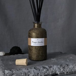 Puede incluir: Un difusor de vidrio dorado con purpurina y varillas negras. La botella tiene una etiqueta que dice "Prague Garden". Un corcho está delante de la botella sobre una superficie de piedra gris.
