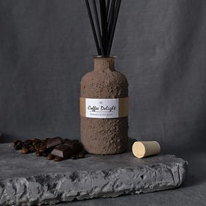 May include: A brown textured diffuser bottle with black reeds, labeled "Coffee Delight." The bottle sits on a gray stone surface with coffee beans, chocolate, and a cork. The background is a dark gray.