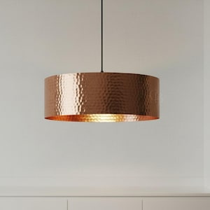 May include: A copper-colored, hammered-metal pendant light fixture. The cylindrical shade hangs from a black cord and a wooden ceiling mount. The light fixture is a warm, metallic color and is designed for indoor use.