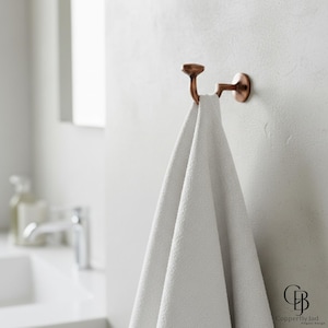 Pode incluir: Um gancho de toalha cor de cobre montado numa parede branca, segurando uma toalha branca texturizada. O gancho tem um design decorativo e faz parte de um conjunto de acessórios de casa de banho. O logótipo "CopperByJad" é visível no canto inferior direito.