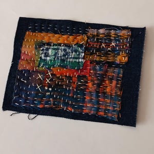 May include: A handcrafted textile art piece with a dark blue denim base. The artwork features patches of colorful fabric in orange, green, and blue, stitched together with white thread. The piece has a rustic, patchwork aesthetic.