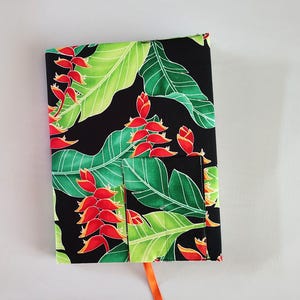 May include: A fabric book cover with a vibrant tropical floral pattern. The cover features large green leaves and red flowers against a black background. An orange ribbon bookmark is visible. The cover has a pocket on the front.