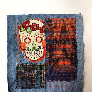 May include: A denim fabric square with a Day of the Dead sugar skull applique. The skull is white with orange accents, red flower crown, and a green mustache. Patches of orange, blue, and green fabric are stitched onto the denim with orange thread.