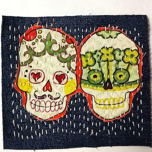 May include: A denim patch featuring two embroidered sugar skull designs. The skulls have colorful floral and heart accents, with orange and yellow outlines. The patch is hand-stitched with white thread.