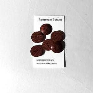 May include: A collection of seven round, dark brown leopard wood buttons, each with two holes. The buttons are 3/4" in diameter and are displayed on a white card that reads "Paramount Buttons" and "LEOPARD WOOD 3/4" Wood from South America."