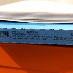 May include: A stack of craft supplies featuring a roll of white fabric atop a blue box labeled "806 STITCH-N-TEAR" with a width of 20 inches. The blue box sits on an orange box.
