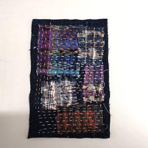 May include: A rectangular textile art piece with a dark blue border. The interior is composed of several patchwork squares in various colors, including purple, blue, orange, and white. The squares are stitched together with visible thread.