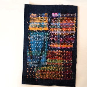 May include: A rectangular textile art piece with a dark blue border. The interior features a patchwork design with vibrant colors including blue, orange, red, and green, stitched together with white thread. The piece has a textured appearance.