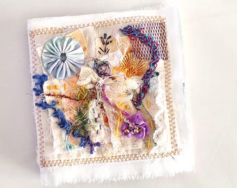 Puede incluir: Una pieza de arte textil cuadrada, de color blanco roto, con una variedad de texturas y colores. Presenta una roseta azul claro, una concha dorada y una flor morada. Los bordes están acabados con una puntada dorada.