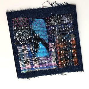 Pode incluir: Uma peça de arte têxtil quadrada com uma borda de ganga azul escura. O centro apresenta vários retalhos de tecido em tons de azul, roxo e laranja, costurados com fio branco. As bordas da ganga estão desfiadas.