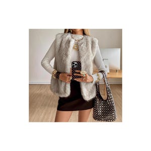 Puede incluir: Un chaleco de piel sintética gris claro se usa sobre una camiseta blanca de manga larga. El atuendo se complementa con joyas doradas, incluyendo un collar, pulseras y anillos. Se sostiene un bolso de mano negro y marrón con tachuelas plateadas.
