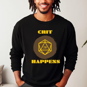 Op de afbeelding: Zwart sweatshirt met de gele tekst "CRIT HAPPENS". Een gele geometrische dobbelsteen met het nummer 20 staat in het midden, tegen een bruine cirkel. Het sweatshirt is een casual kledingstuk.