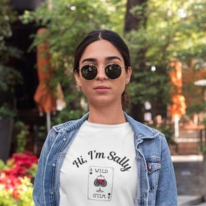 May include: A woman wearing a white t-shirt with the text "Hi, I'm Sally" and a playing card design. She is also wearing a denim jacket and round sunglasses. The background is blurred with greenery.