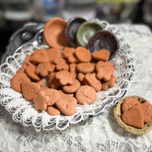 May include: A collection of terracotta-colored clay shapes, including circles, flowers, and hearts, are displayed in a decorative silver tray. Several small ceramic bowls in various colors are also present. The items are arranged on a white lace tablecloth.