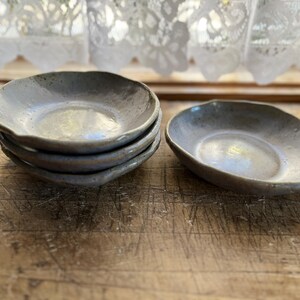 May include: A stack of four small, handmade ceramic plates with a light gray glaze and irregular, organic shapes. One additional plate sits beside the stack. The plates are on a wooden surface, with a lace curtain in the background.