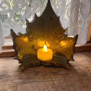 May include: A decorative, leaf-shaped candle holder in a metallic gold finish. The holder cradles a small, round, yellow LED candle, creating a warm glow. The candle holder is placed on a wooden surface.