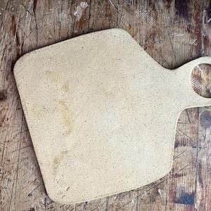 May include: A speckled, beige ceramic cutting board with a handle. The board has a unique shape with a rounded handle and a textured surface. The cutting board is approximately 12 inches long and 9 inches wide.