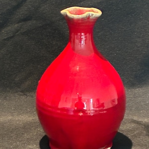 May include: A glossy red ceramic vase with a bulbous body and a slender neck. The vase has a ruffled rim and sits on a black coaster. The vase is approximately 8 inches tall.