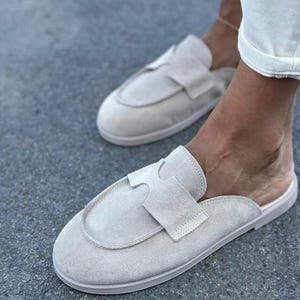 May include: Light beige suede slip-on shoes with a rounded toe and a decorative strap across the top. The shoes have a low profile and a neutral colour, suitable for casual wear. The soles are a matching light beige colour.
