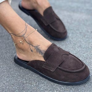 May include: Brown suede slip-on loafers with a black sole. The shoes have a classic design with a strap across the top. The image shows a pair of the shoes, with one shoe in focus.